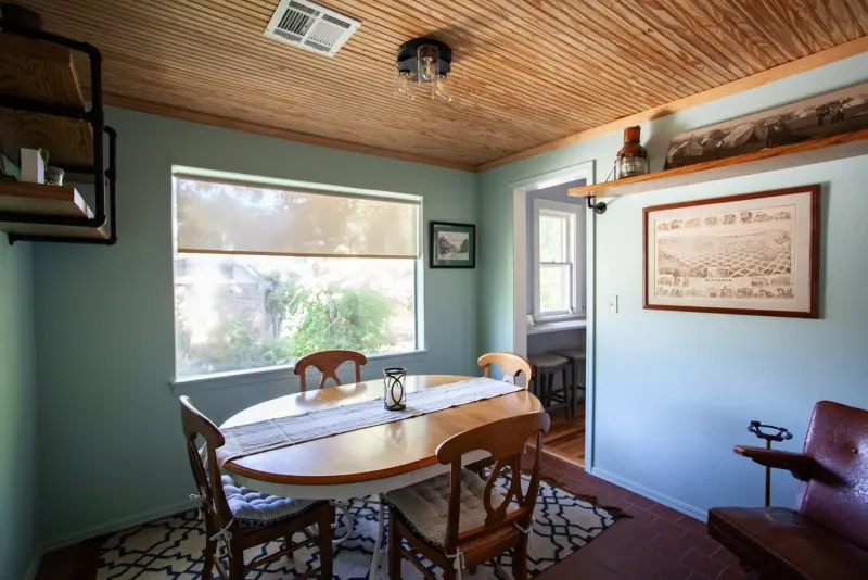 Breakfast room with a round wooden table and chairs, a large window and a wood-plank ceiling