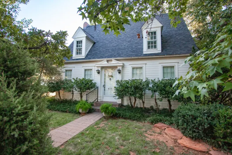 The white Cape Cod-style cottage with two dormer windows, behind a stone path, shrubs and tall trees