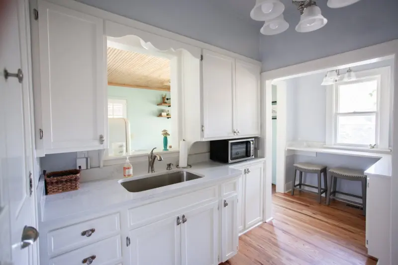 Kitchen sink under a window, white cabinets and a microwave, with a breakfast bar beyond